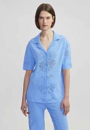 Chemise à manches courtes bleu clair, à boutons, en tissu maillé texturé avec un motif de feuilles. Le col est plat, offrant une coupe décontractée.