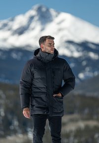 Black puffer jacket with a high collar and hood, featuring zippered pockets and a smooth texture, worn over dark pants against a snowy mountain backdrop.