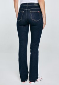 Osoba u tamnoplavim trapericama širokih nogavica i bijelim tenisicama, prikazana s leđa na jednostavnoj svijetloj pozadini.