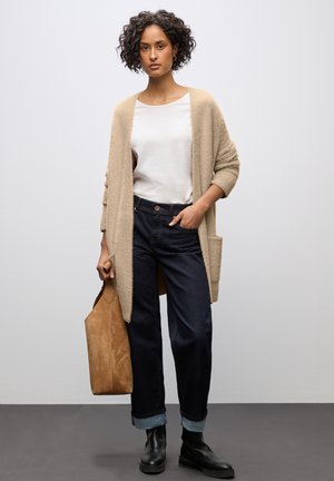 Beige fuzzy cardigan over een wit top, donkere denim wijdgeknoopte jeans, zwarte enkellaarsjes en een lichtbruine suède tas. Eenvoudig ontwerp en texturen.