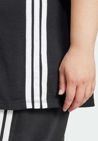 Melna kokvilnas sporta apģērba daļa ar trim vertikālām baltām svītrām sānos, ar tuvplānu, kurā redz roka, kas atpūšas uz auduma.