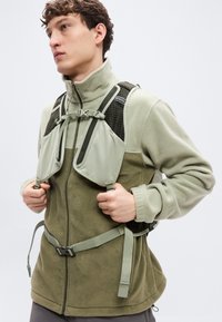 Ζακέτα από φλίς σε ανοιχτό και σκούρο ελιά με ψηλό γιακά, ρυθμιζόμενες λωρίδες και υφασμάτινη επιφάνεια στους ώμους.