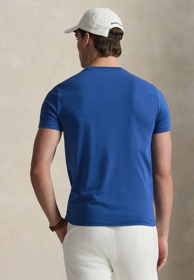 Μπλε κοντομάνικη μπλούζα από λείο βαμβακερό ύφασμα, με κλασικό στρογγυλό λαιμό και εφαρμοστή γραμμή, φορεμένη με λευκό παντελόνι και καπέλο.