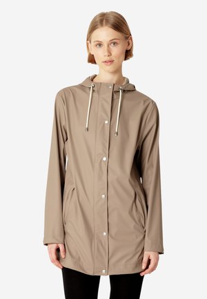 Młoda kobieta ubrana w jasnobrązowy płaszcz przeciwdeszczowy z kapturem, zatrzaskami i przednimi kieszeniami, stojąca na jednolitym tle.