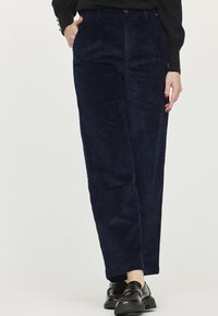 Pantalons en velours côtelé bleu marine avec une coupe droite, dotés de poches latérales et d'une fermeture à bouton, associés à des chaussures en cuir noir.