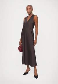Femme en robe midi sans manches marron foncé, tenant une pochette bordeaux, portant des talons noirs et de grandes boucles d'oreilles blanches en anneau, se tenant devant un fond uni.