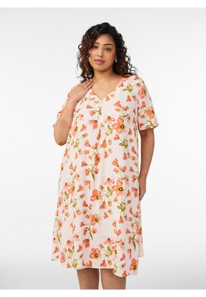 Frau mit lockigem Haar, die ein knielanges weißes Kleid mit rosa Blumenmuster trägt und vor einem schlichten hellen Hintergrund steht.