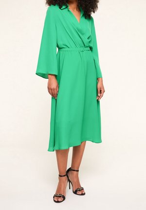Green wrap dress with a belted waist, long bell sleeves, and a flowing hem. Paired with black high-heeled sandals. Smooth texture.