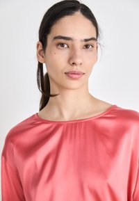Rosa satengtop med løs passform, vid utringning og lange ermer. Glatt tekstur med subtil glans, båret av en person mot en ensfarget bakgrunn.