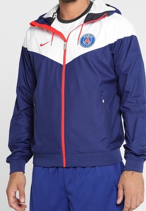 Mężczyzna ubrany w niebiesko-białą kurtkę Paris Saint-Germain z czerwonym suwakiem i logo Nike, w połączeniu z niebieskimi spodniami, stojący na jednolitym tle.