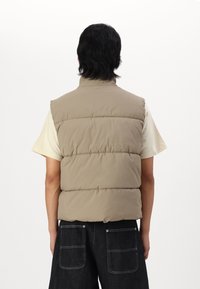 Tan puffer vest with a high collar, quilted design, and sleeveless cut, worn over a light-colored t-shirt and paired with dark jeans.