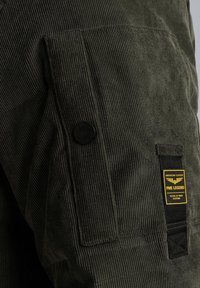 Donkergroene corduroy jas met een gestructureerd oppervlak, voorzien van een zijzak, een zwarte knoop en een gele merketiket.