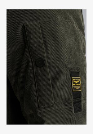 Dunkelgrüne Cordjacke mit einer strukturierten Oberfläche, ausgestattet mit einer seitlichen Tasche, einem schwarzen Knopf und einem gelben Markenetikett.