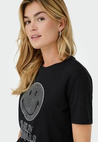 T-shirt en coton noir présentant un grand graphique de smiley blanc avec un design texturé et le texte "Smiley Originals" en gras.
