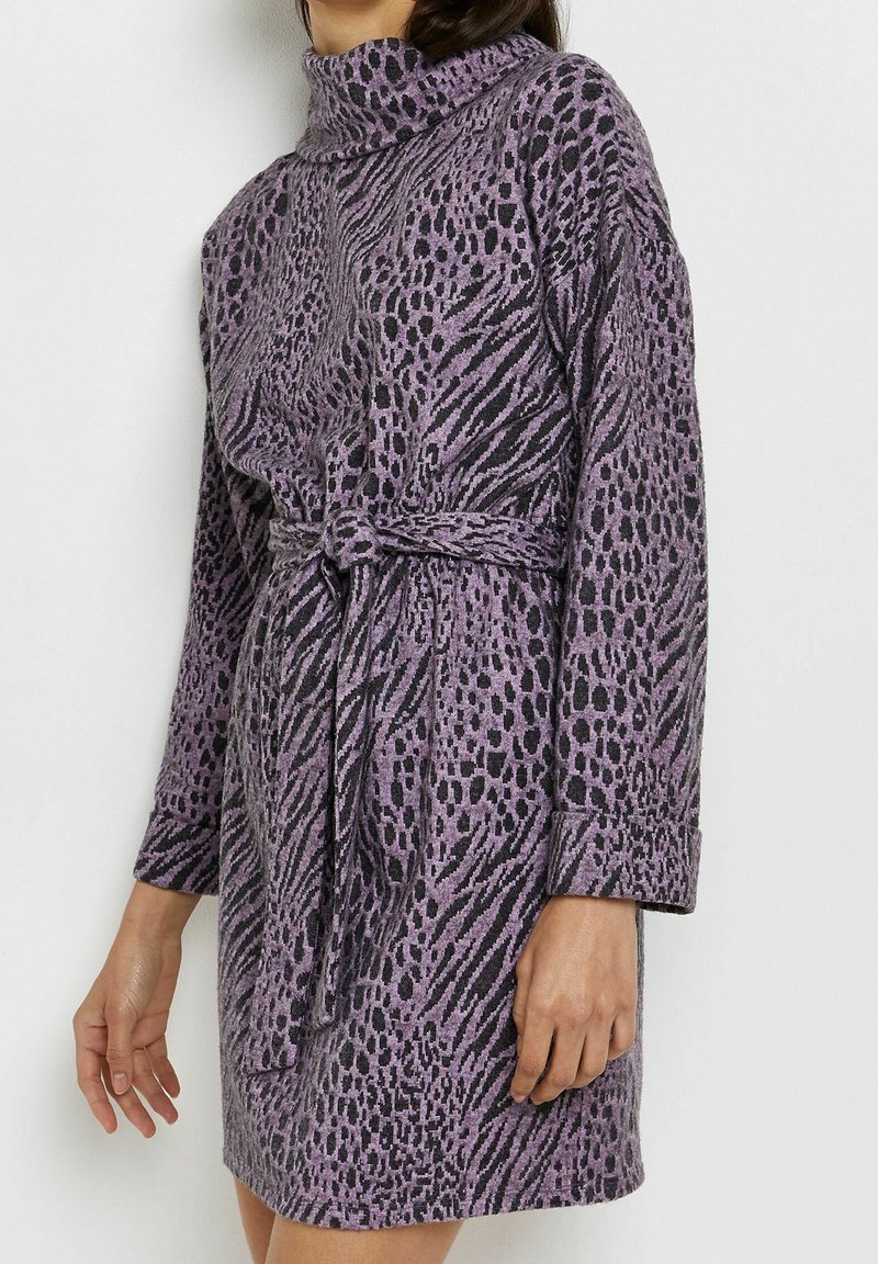 Femme portant une robe à manches longues imprimée animalier violet et noir, avec un col roulé et une ceinture nouée à la taille.