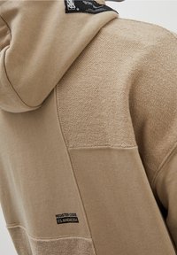 Sweat à capuche beige avec un tissu texturé, présentant un patch au niveau du col et divers motifs de couture sur les manches et le corps.
