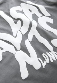 Białe, pogrubione, nieregularne litery tworzące "ASAI AN" i częściowo "LONDON" na szarym tle materiału.