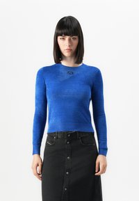 Vrouw met steil zwart haar in een getailleerd blauw langemouwen top en een hooggesneden zwarte knopen-denimrok staat tegen een witte achtergrond.