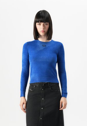 Vrouw met steil zwart haar in een getailleerd blauw langemouwen top en een hooggesneden zwarte knopen-denimrok staat tegen een witte achtergrond.