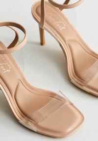Kroppsnära högklackade sandaler med ankelslinga i nude-färgat läder, genomskinlig tåslinga, fyrkantig tåform och slät yta. Texturerad innersula för ökad komfort.