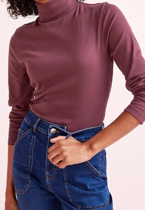 Osoba nosząca dopasowany, prążkowany, mauve'owy golf z długimi rękawami oraz wysokimi niebieskimi dżinsami z przeszyciami i zapięciem na guzik.