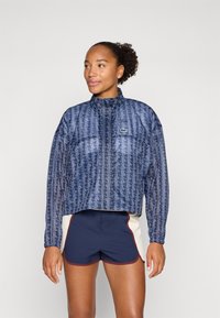 Navy patterned windbreaker with a high collar and elastic cuffs, worn over navy and cream shorts with red accents. Features brand logo.