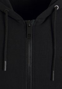 Felpa nera con zip, caratterizzata da un tessuto liscio, cordini e una robusta cerniera in plastica con un motivo geometrico sulla linguetta.