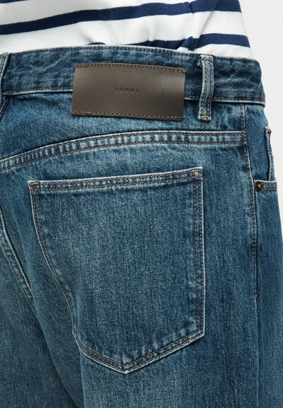 Μπλε τζιν τζιν με ελαφριά ξεθώριασμα, με καφέ δερμάτινο ταμπελάκι στη μέση, ραφές στις πίσω τσέπες και κλασικό σχέδιο με πέντε τσέπες.