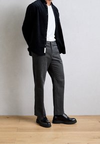 Camicia di velluto a coste nera, t-shirt bianca sotto, pantaloni grigi traforati e scarpe nere con lacci, in piedi su pavimento di legno chiaro.