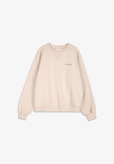Sweat-shirt beige clair à col rond avec manches longues bouffantes et petit texte "HUG ME" sur la poitrine gauche.
