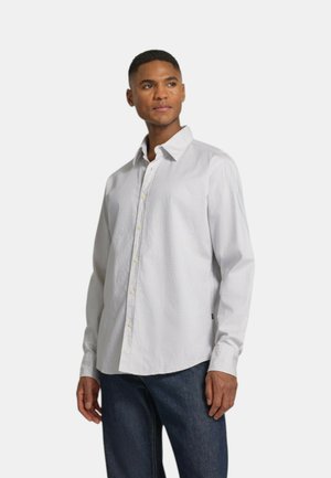 Mann, der ein weißes langärmeliges Hemd und dunkelblaue Jeans trägt, steht und schaut zur Seite vor einem schlichten Hintergrund.