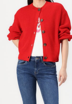 Kvinde iført en knaldrød knaplukket strikcardigan med opkrogede ærmer over en hvid t-shirt med tryk og blå jeans.