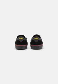 Melnas zamšādas sporta apavus ar plakanu zoli, dzeltenu "etnies" logo uz papēža un sarkaniem akcentiem ap apakšējiem maliem. Strukturēta virsma lielākai saķerei.