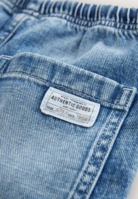 Blåt denimstof med et hvidt rektangulært mærke med teksten "Authentic Goods" syet nær en lomme-søm.