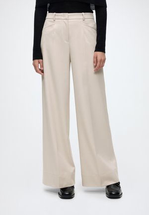 Persona che indossa pantaloni beige a gamba larga, top nero a manica lunga e scarpe nere, in piedi su uno sfondo bianco uniforme.
