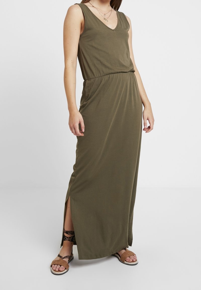 Oliwkowa maxi sukienka z dekoltem w serek, bez rękawów, z elastyczną talią i rozcięciami po bokach. Zestawiona z brązowymi płaskimi sandałami.