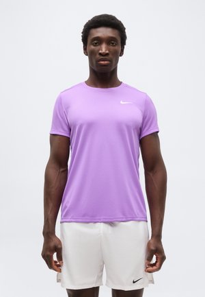 Egy férfi áll előre nézve, egyszínű lila Nike pólót és fehér Nike rövidnadrágot visel, semleges háttér előtt.