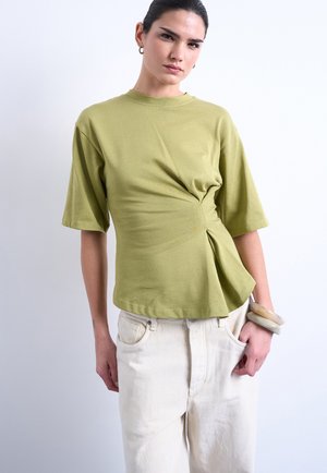 Γυναίκα που φοράει πράσινο ελιάς κοντομάνικο τοπ με πλάγια στροφή, εκρού παντελόνια ψηλόμεσα και μεγάλα βραχιόλια σε χρώμα πέτρας στον δεξί καρπό.