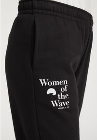 Sorte joggere med elastisk midje, med hvit tekst "Women of the Wave" og logo på siden. Myk stoff, vanlig passform.