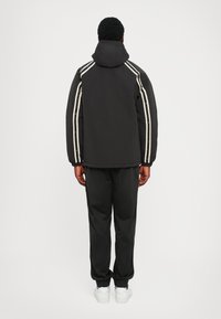 Črna prešita jakna z belimi črtami ob straneh in kapuco, kombinirana s črnimi hlačami in belimi supergami. Pogled od zadaj poudarja dizajn in prileganje.