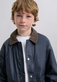 Niño pequeño con cabello castaño claro y pecas lleva una chaqueta oscura con un cuello de pana marrón sobre una camisa blanca, contra un fondo liso.