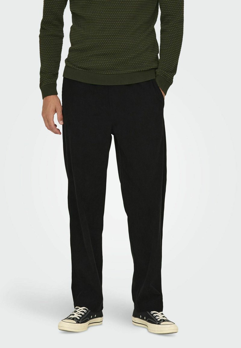 Schwarze Corduroyhosen mit lässiger Passform, seitlichen Taschen und geripptem Bündchen. Kombiniert mit schwarzen Turnschuhen und einem strukturierten grünen Pullover.
