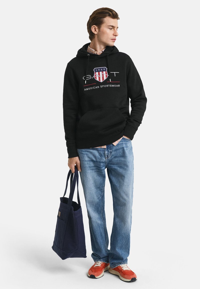 Svart huvtröja med logotyp på framsidan, blå jeans, röda och beige sneakers, samt en marinblå tygväska med handtag. Avslappnad outfit.