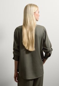 Frau mit langen, glatten blonden Haaren, die ein olivgrünes, langärmliges Shirt und eine Hose trägt, steht mit dem Rücken zu einer schlichten weißen Wand.