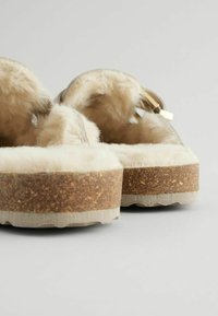 Bruna tofflor med korksula, klädda med en mjuk, krämfärgad konstpäls foder och en mjuk, lätt strukturerad ovandel. Har en rundad form och minimal accent.