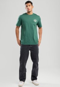 T-shirt verde in cotone con maniche corte e scollatura rotonda, caratterizzato da un design grafico bianco sul lato sinistro del petto. Indossato con jeans neri.