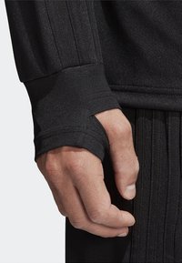 Manguito de una camiseta deportiva negra de manga larga con orificio para el pulgar y tela texturizada. Primer plano de una mano sujetando la tela, mostrando los detalles de las costuras.