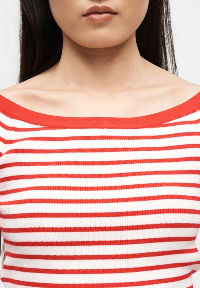 Γυναίκα με μακριά σκούρα μαλλιά, που φοράει λευκή μπλούζα εκτός ώμων με κόκκινες οριζόντιες ρίγες, εμφανίζοντας τον λαιμό και τις κλείδες.