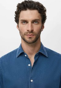 Mann mit kurzen lockigen braunen Haaren und Bart, der ein blaues Jeanshemd mit Knöpfen trägt und frontal vor einem weißen Hintergrund steht.
