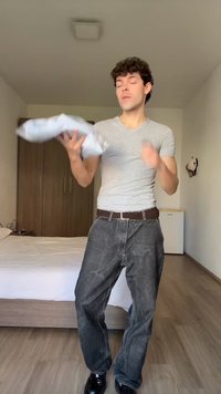 Jauns vīrietis pelēkā kreklā un džinsos met spilvenu guļamistabā ar koka drēbju skapi un gultu fonā.